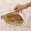A hand is opening a white fabric storage bag with a zipper to reveal a beige interior holding makeup brushes.