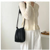 A woman is holding a black pleated PU leather crossbody bag while wearing a cream knit top and white trousers.