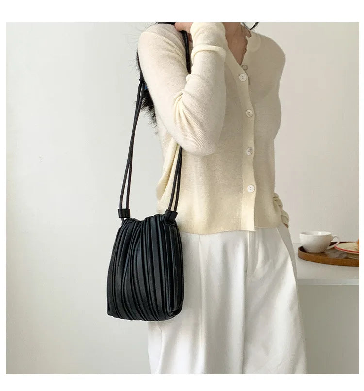 A woman is holding a black pleated PU leather crossbody bag while wearing a cream knit top and white trousers.