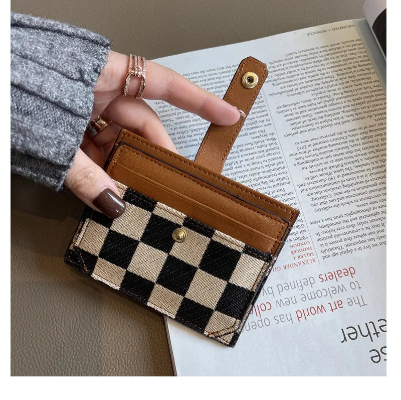 A hand holds a brown checkered PU leather mini card wallet with a leather strap against a background of printed text.
