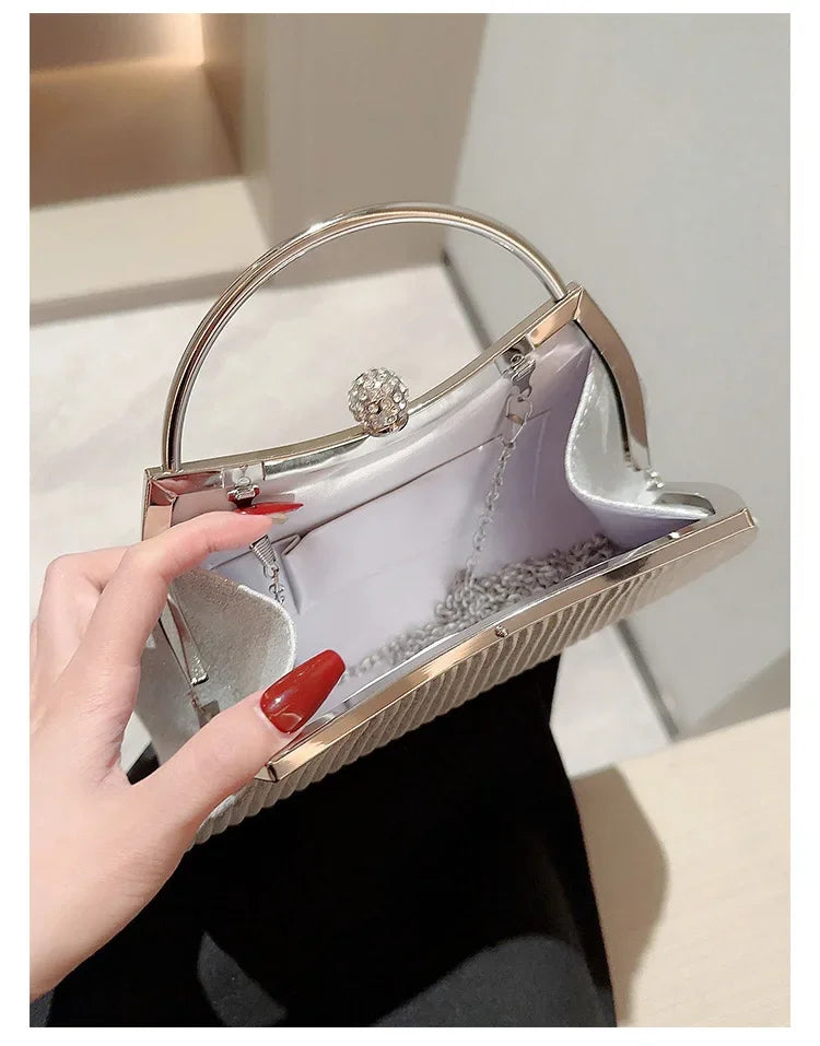 A hand holds a silver pleated glitter evening handbag with a top handle and chain strap.