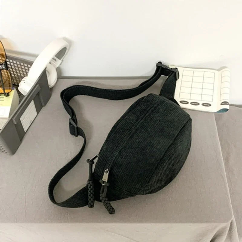 A black corduroy waist pack for women is displayed on a gray surface next to a white notepad.