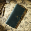 A dark green genuine leather long bi-fold wallet with a brass snap closure resting on aged parchment paper.