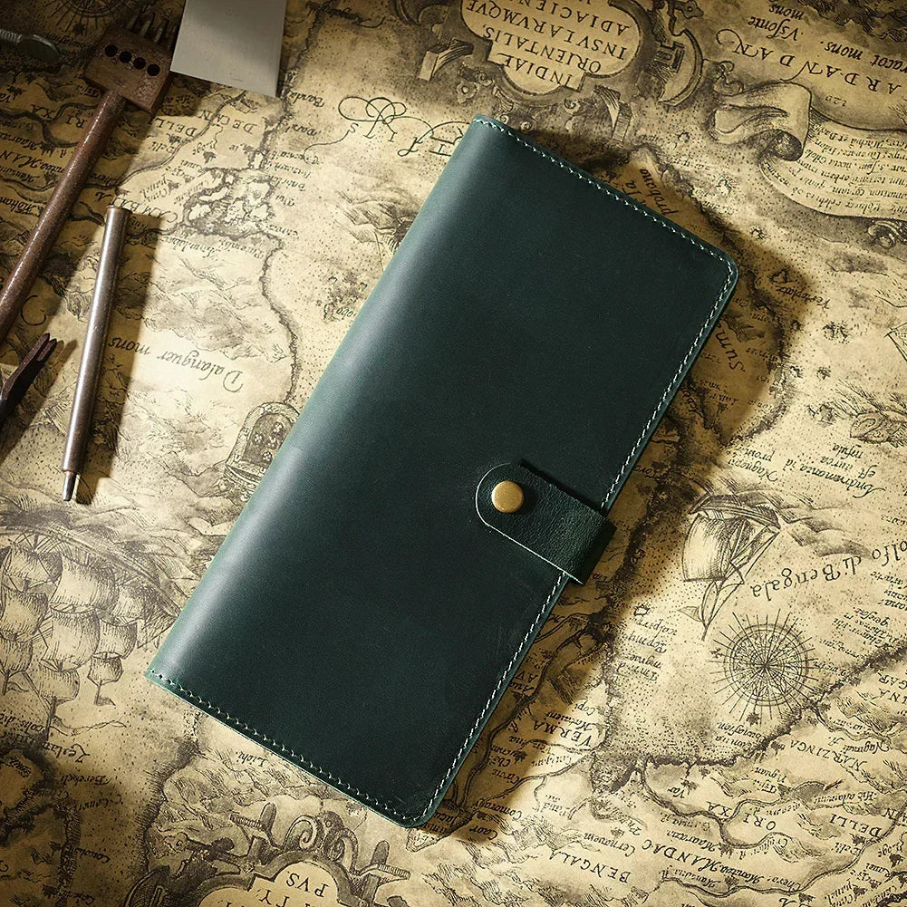 A dark green genuine leather long bi-fold wallet with a brass snap closure resting on aged parchment paper.