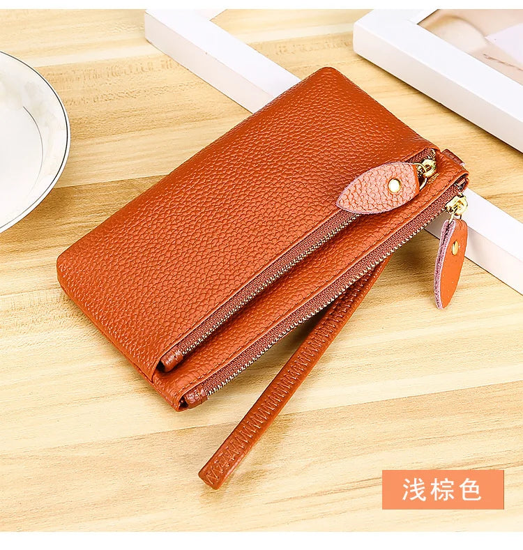 A brown genuine leather wristlet clutch wallet with a zipper closure is shown on a wooden surface.