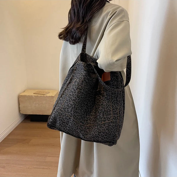Eine Frau hält eine dunkle Leopardenmuster-Vintage-Canvas-Schultertasche vor einem hellen Kleidungsstück.