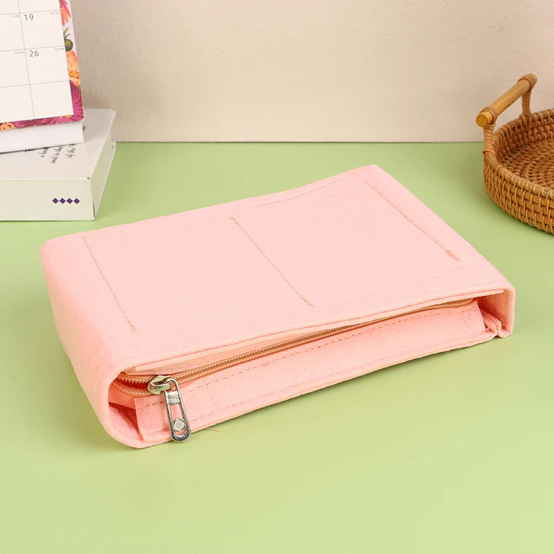A pink felt cosmetic bag insert organizer lying on a green surface next to a woven basket and some boxed items.