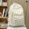 Mochila blanca con estampado floral y múltiples bolsillos, apoyada sobre una superficie clara junto a libros.