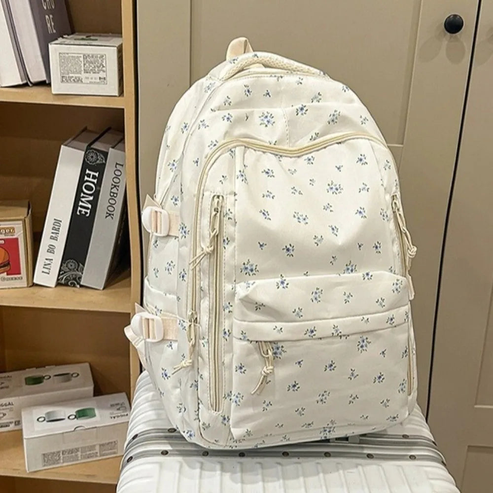 Mochila blanca con estampado floral y múltiples bolsillos, apoyada sobre una superficie clara junto a libros.