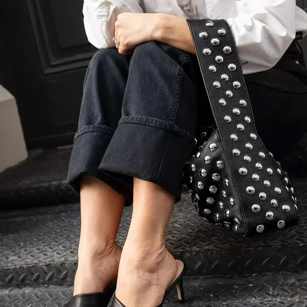 A close-up of a black leather shoulder bag adorned with silver metal rivets, resting next to a person wearing dark jeans.