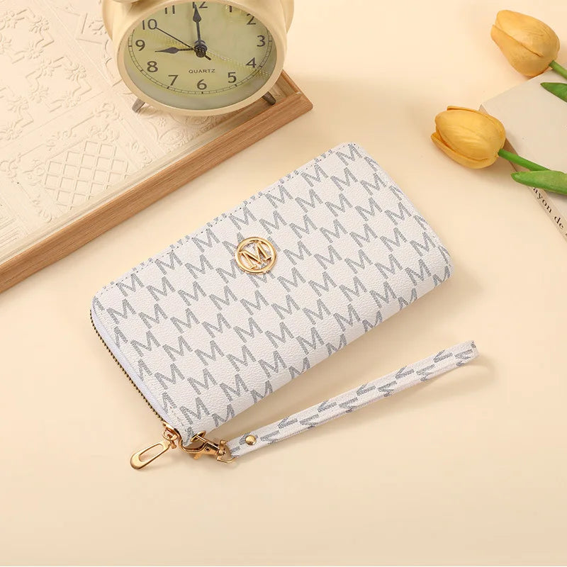 A light gray zippered wallet with a gold logo is displayed next to a vintage-style clock and yellow tulips.