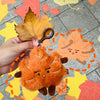 An orange maple leaf plush keychain charm is being held against a background of colorful autumn leaves.