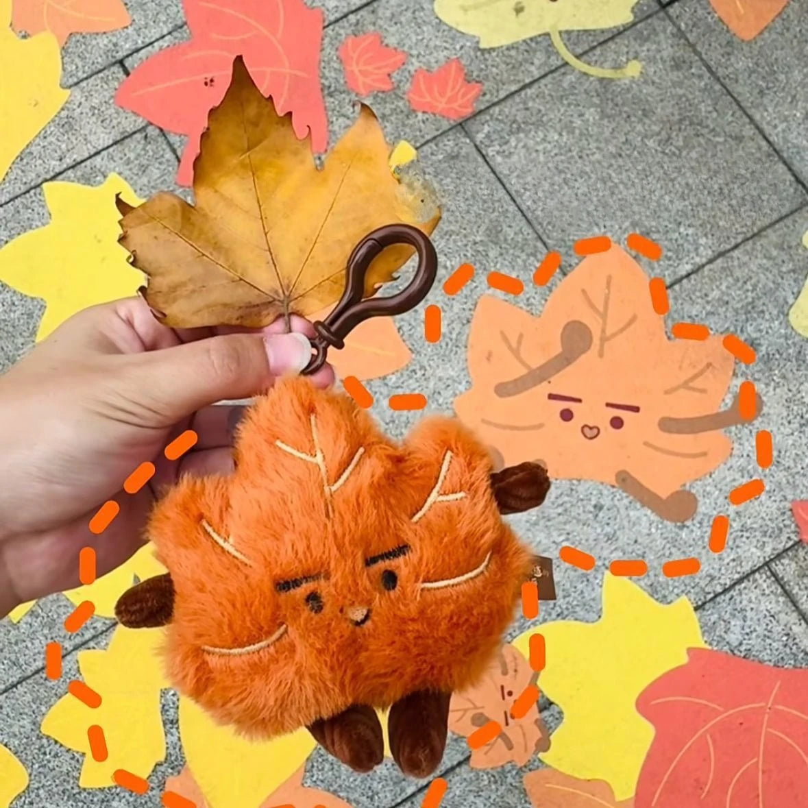 An orange maple leaf plush keychain charm is being held against a background of colorful autumn leaves.
