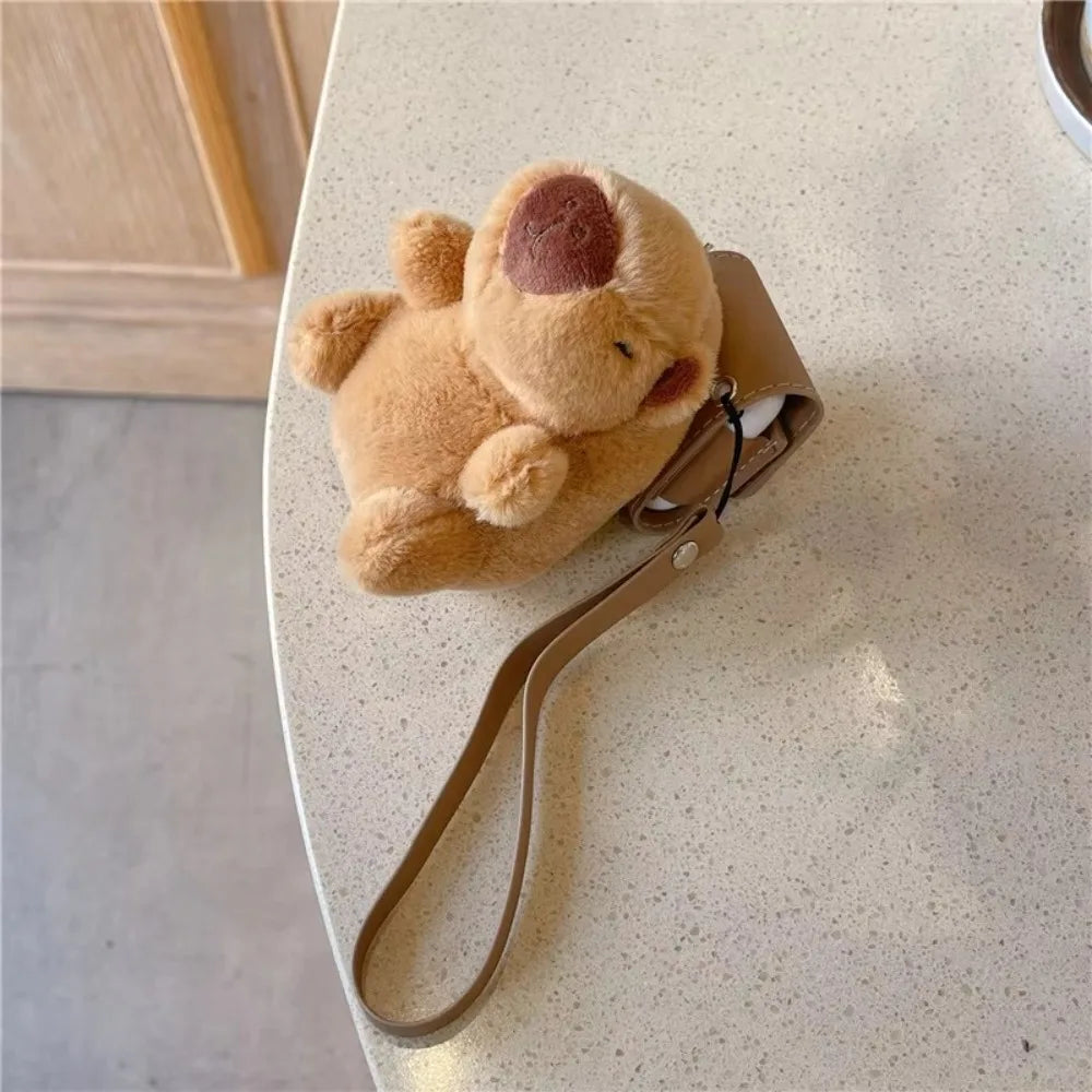 A fluffy brown elephant plush toy is shown resting in a tan leather case for AirPods on a light-colored table.