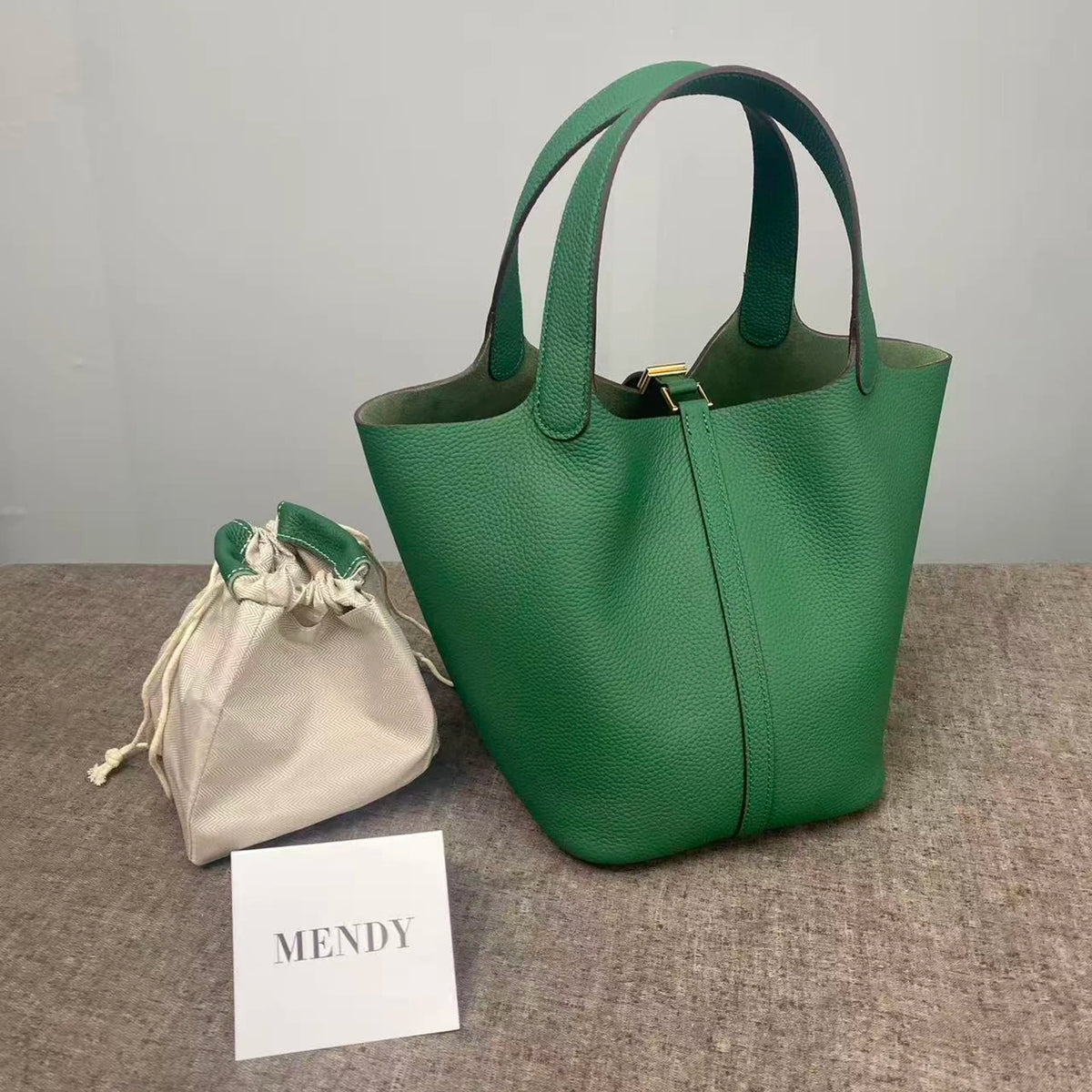 A dark green leather bucket bag is displayed next to a light beige drawstring pouch and a card labeled MENDY.