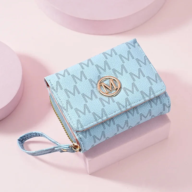 A light blue elegant letter zipper wristlet wallet with a gold monogram detail resting on a pink surface.