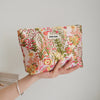 A hand holds a floral canvas makeup bag featuring pink, yellow, and green botanical prints.