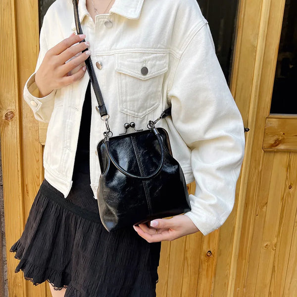 A woman holds a black leather shell handbag with a strap against a light jacket and a black skirt.