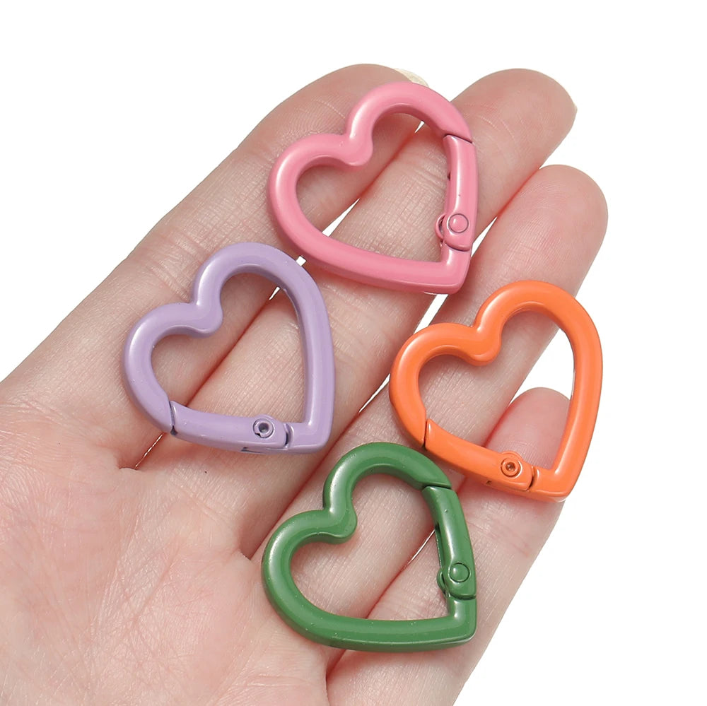 A hand holds four heart-shaped metal spring clasps in pink, purple, orange, and green.