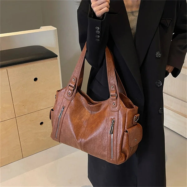 A brown leather tote bag with a multi-pocket design is held by a person wearing a black coat.