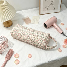 Una bolsa de costura floral rosa reposa sobre un paño blanco junto a un secador y un peine rosa.