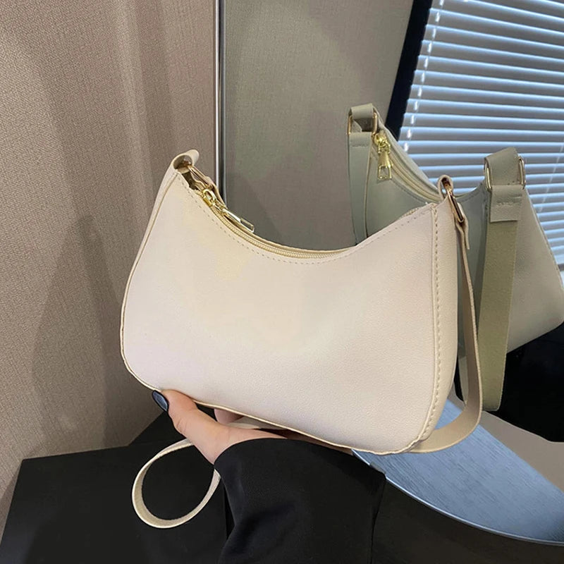 A light beige leather shoulder bag with a gold zipper and strap is held by a hand against a blurred background.