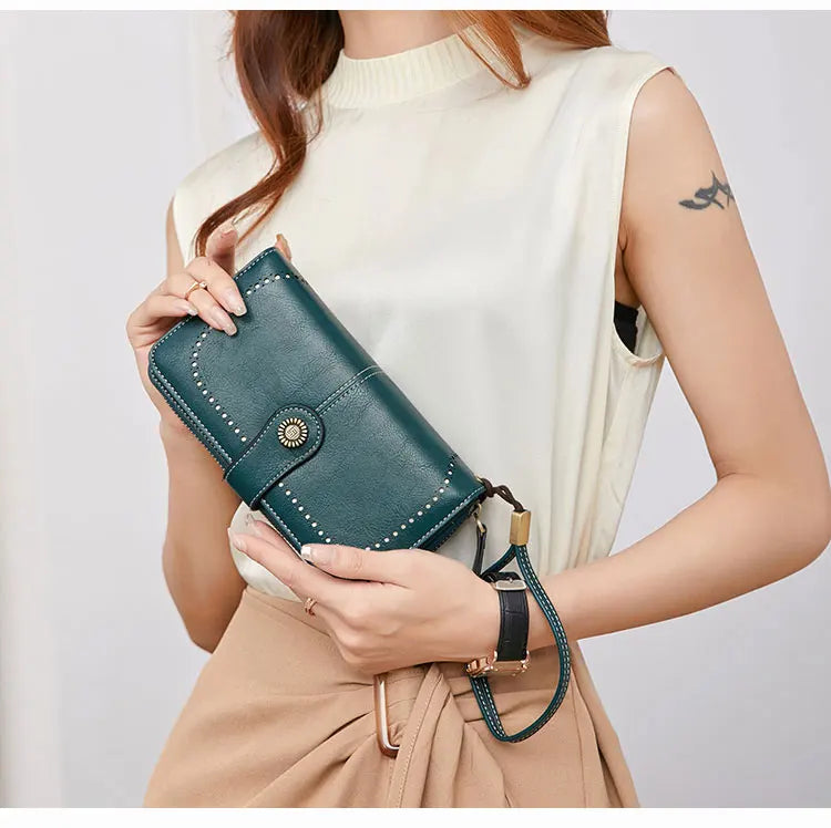 A woman holds a dark teal RFID anti-theft multi-card wallet with a leather wrist strap against a cream top and beige skirt.