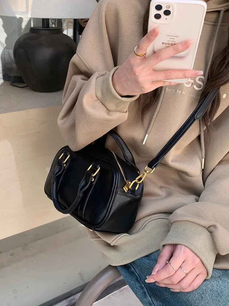 A person holds a black vintage PU leather shoulder bag with gold hardware against a light brown hoodie.