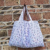 A white reusable shopping bag with a brown strap is displayed against a brick wall.