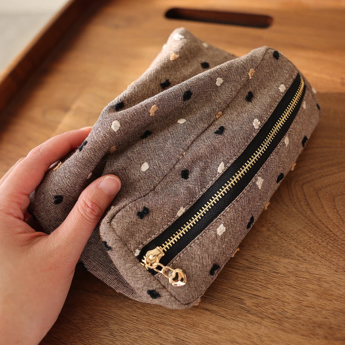 Une main tient une pochette de rangement cosmétique en tissu brun à pois blancs et une fermeture éclair noire sur une surface