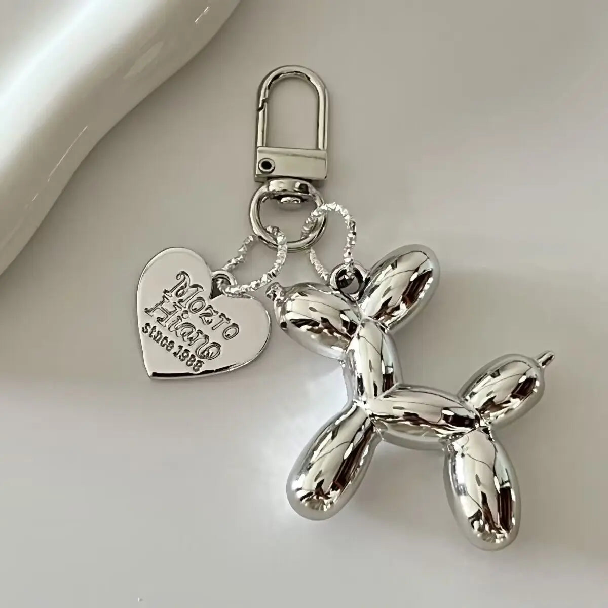 Silver balloon dog keychain charm with a heart tag and jump ring on a white surface