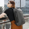 A woman wearing a black shirt and brown pants is holding a gray anti-theft waterproof laptop backpack.