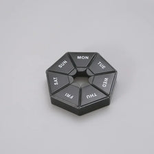 A black, hexagonal weekly pill organizer divided into seven sections labeled with the days of the week.