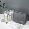 Sac organisateur en tissu gris avec des produits de soin et de beauté, incluant des flacons et une serviette blanche.