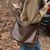 A person is holding a brown vintage PU leather tote bag with a gold crossbody strap outdoors on a leaf-covered ground.