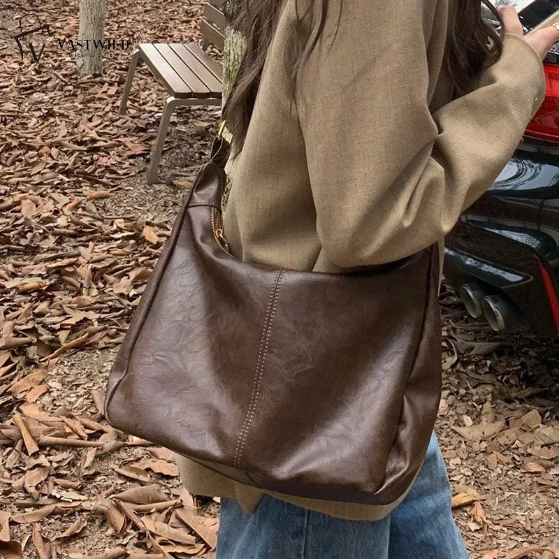 A person is holding a brown vintage PU leather tote bag with a gold crossbody strap outdoors on a leaf-covered ground.