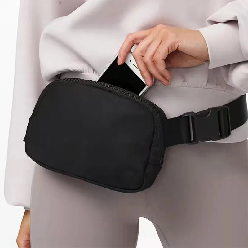 A woman wearing a light jacket holds a black waist bag across her torso.
