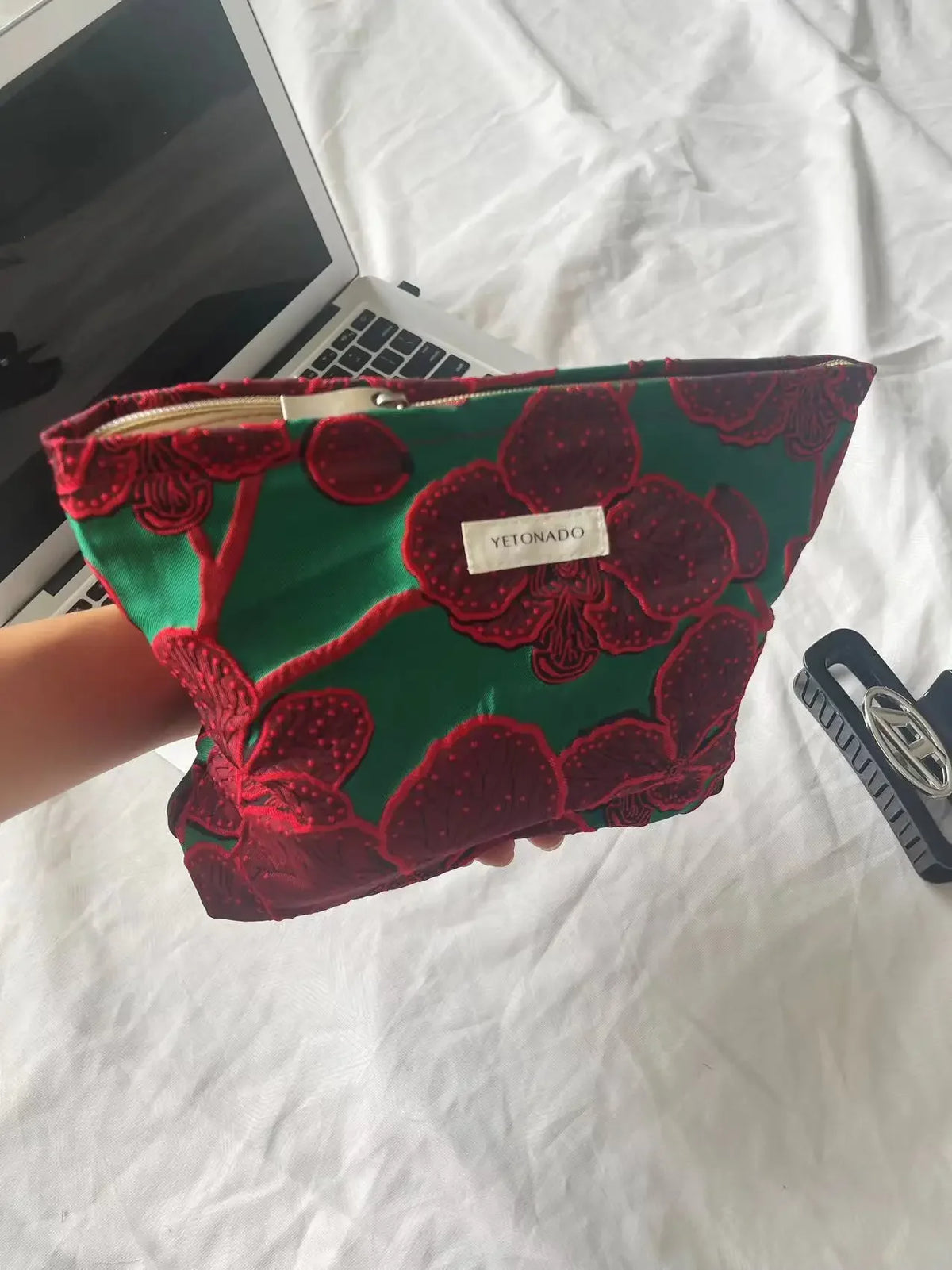 A vibrant green velvet cosmetic pouch with red floral embroidery and a zipper is held against a white background.