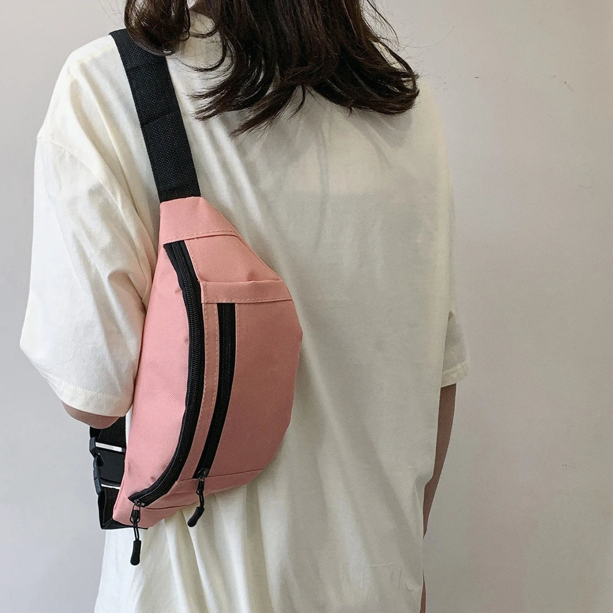 A pink nylon waist bag with black straps is worn on a white t-shirt.