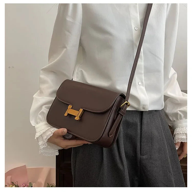 Eine Person hält eine braune Leder-Ein-Schultertasche vor einem weißen Hemd und einem grauen Rock.