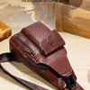 A brown genuine leather sling backpack is shown lying on a wooden surface with a leather strap.