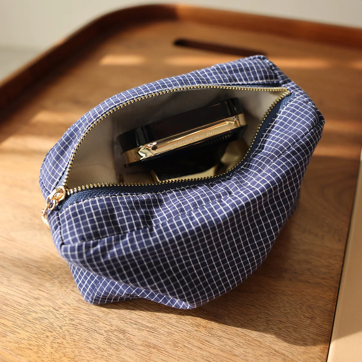 Pochette à motif à carreaux bleu marine avec fermeture éclair dorée contenant un produit cosmétique noir sur une surface en b