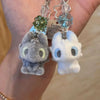 A pair of Toothless cartoon plush keychains with green and blue crystal charms hanging from a silver chain.