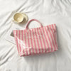 A pink and white striped transparent PVC tote bag is displayed on a white sheet next to a cup.