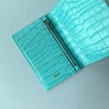A turquoise crocodile pattern leather passport holder with a silver zipper and the initials B.O.