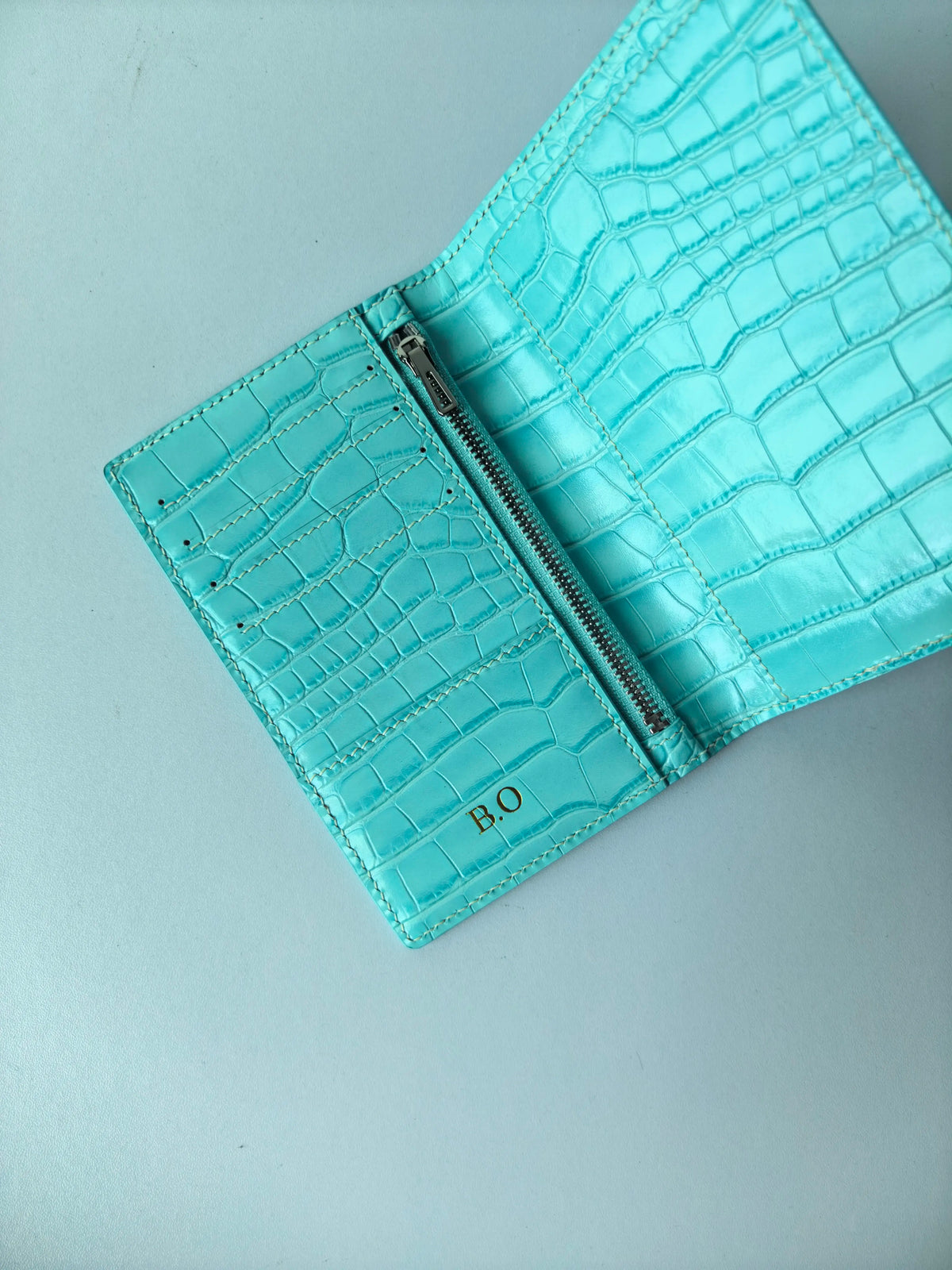 A turquoise crocodile pattern leather passport holder with a silver zipper and the initials B.O.