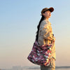 A person wearing a beige canvas outfit and hat stands by water, holding a pink camouflage shoulder bag.
