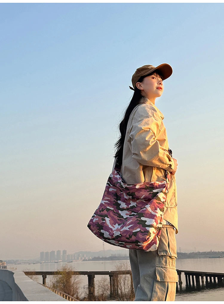 A person wearing a beige canvas outfit and hat stands by water, holding a pink camouflage shoulder bag.