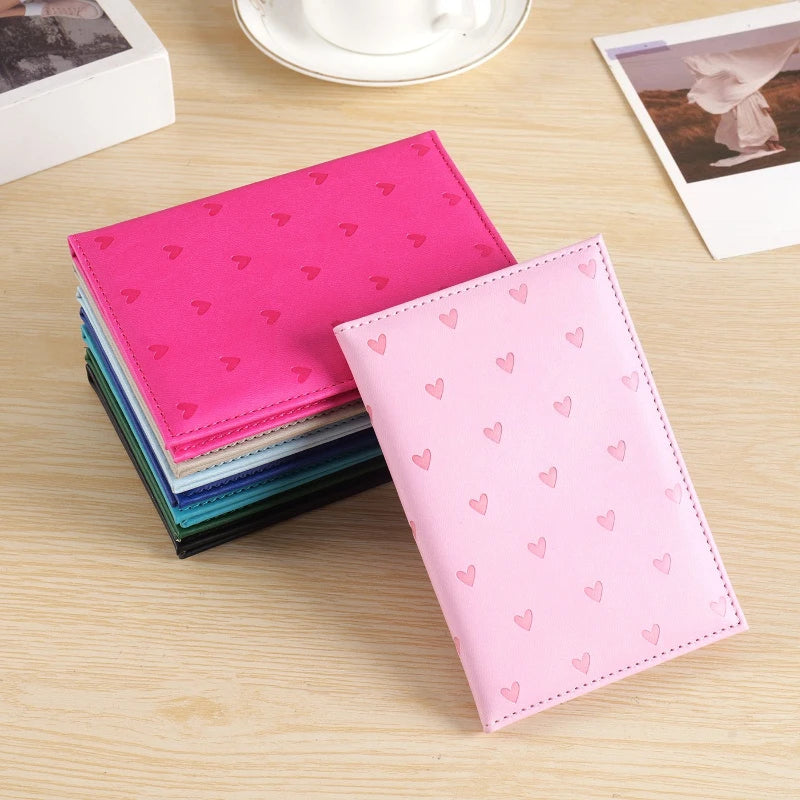 A stack of pink and teal heart print passport covers and a single pink heart print wallet on a wooden surface.