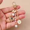 A gold-toned pearl bow heart keychain with a dangling charm is held in a person's hand.